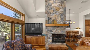 A spacious living room with a stone fireplace, a large flat-screen TV, wooden furniture, and a view of the outdoors. - Legends - Simply Perfect Elkhorn Retreat (Sun Valley)