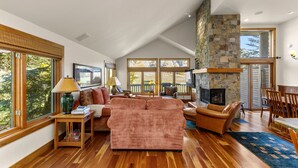 A spacious living room with a stone fireplace, wooden flooring, and large windows offering a view of the outdoors. - Legends - Simply Perfect Elkhorn Retreat (Sun Valley)