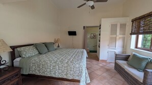 A bedroom with a bed, a nightstand, a lamp, a ceiling fan, a window with blinds, and a door leading to a bathroom. - Charming 1-bedroom apartment at La Ropa Beach (Zihuatanejo)