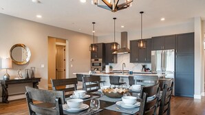 A modern kitchen with dark cabinets, stainless steel appliances, pendant lighting, and a dining area with wooden chairs and a central island.