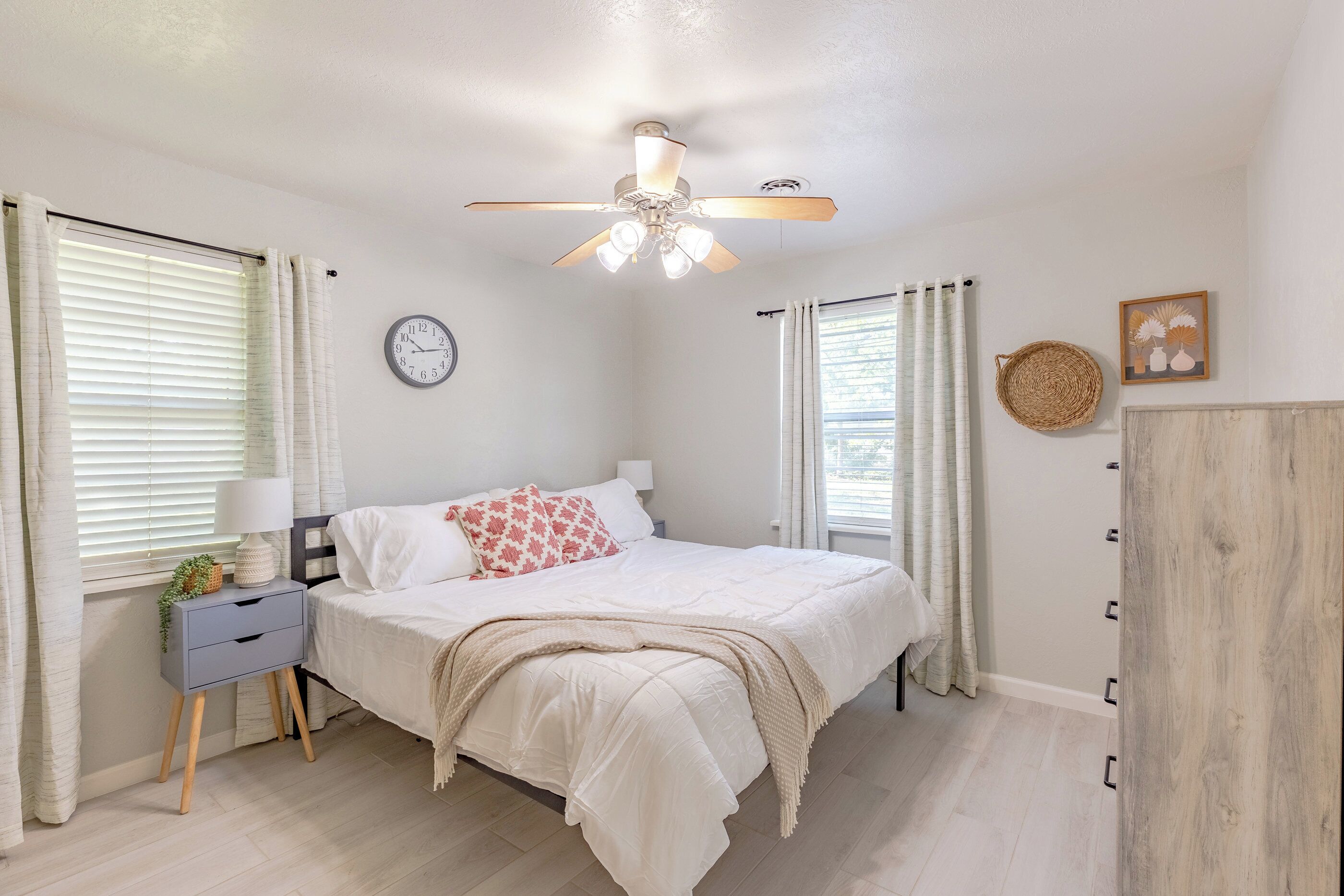 A bedroom with a bed, bedside table, lamp, clock, ceiling fan, and curtains.