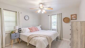 A bedroom with a bed, bedside table, lamp, clock, ceiling fan, and curtains.