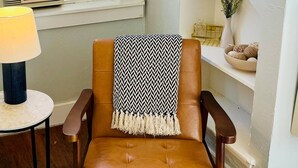 A brown leather chair with a zigzag patterned throw blanket, a round table with a lamp, and a window with curtains. - Carriage House studio min to ECU (Ada)