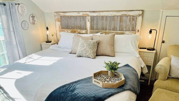 A large bed with a wooden headboard, a bedside table with a lamp, and a chair. - Carriage House studio min to ECU (Ada)