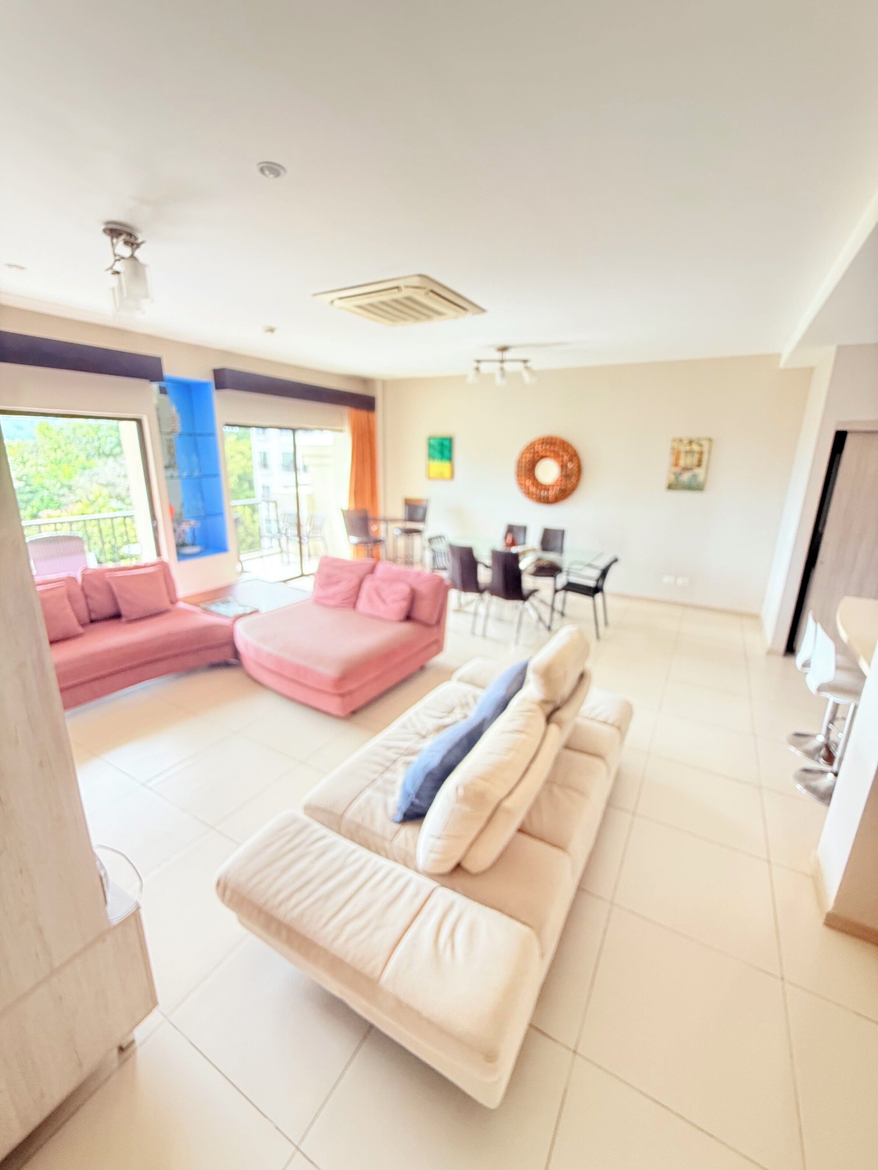Un'ampia zona giorno con un divano color beige, un divano rosa e un angolo chaise longue. C'è un'area pranzo con sedie e un tavolo. La stanza ha ampie finestre e una ventola a soffitto.