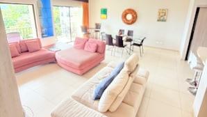 Ein großzügiger Wohnbereich mit einem beigen Sofa, einem rosa Sofa und einer Liege. Es gibt einen Essbereich mit Stühlen und einem Tisch. Der Raum verfügt über große Fenster und einen Deckenventilator.