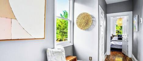 Un couloir avec un banc, une décoration murale ronde en fibres tressées et un lit visible à travers une porte ouverte.
