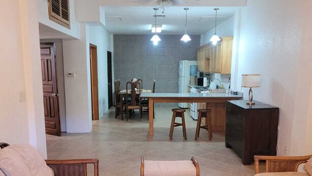 A spacious living room with a sofa, a wicker coffee table, and a dining area with a table and chairs. A kitchen with a refrigerator and cabinets is visible in the background.