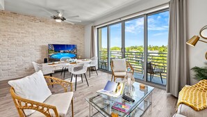 A spacious living area with a glass coffee table, wicker chairs, a television, and a view of a swimming pool. - Waterfront Condo with Pool, Spa, and Fitness Center near Downtown Sarasota (Sarasota)
