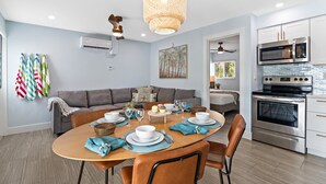 A modern kitchen with a stainless steel oven and microwave, a dining area with a round table and chairs, and a gray sofa in the background.