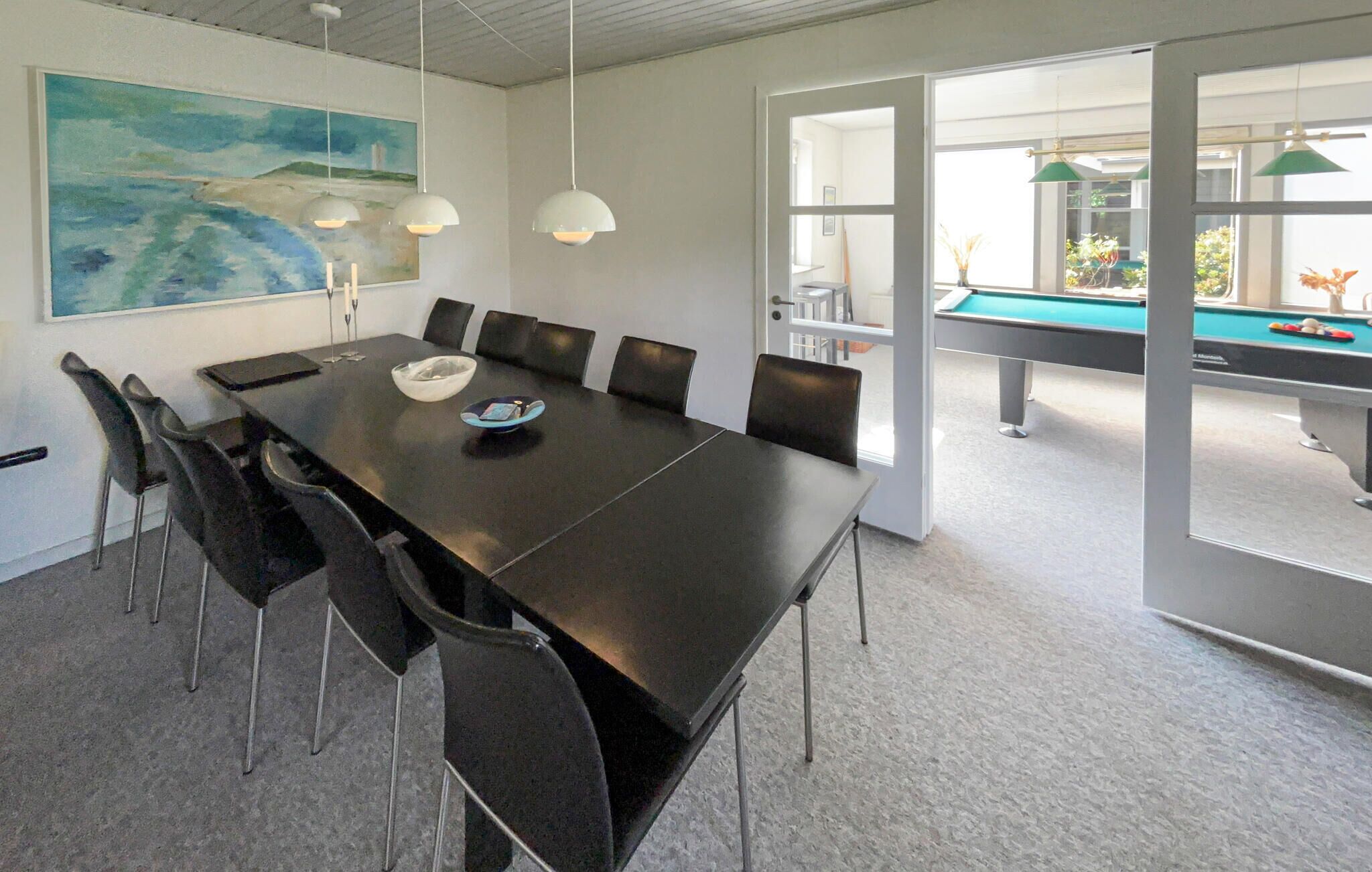 A modern conference room with a long table, chairs, a painting, pendant lights, and a pool table visible through an open door.
