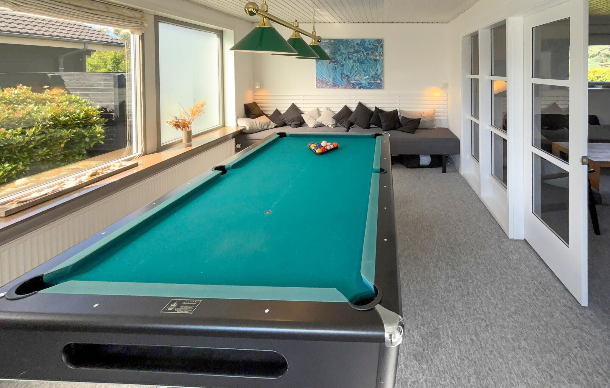 Een pooltafel met een groene viltbekleding, een set poolkeu's en een rack biljartballen. Er is een bank met kussens, een raam met uitzicht op groen, en een schilderij aan de muur.