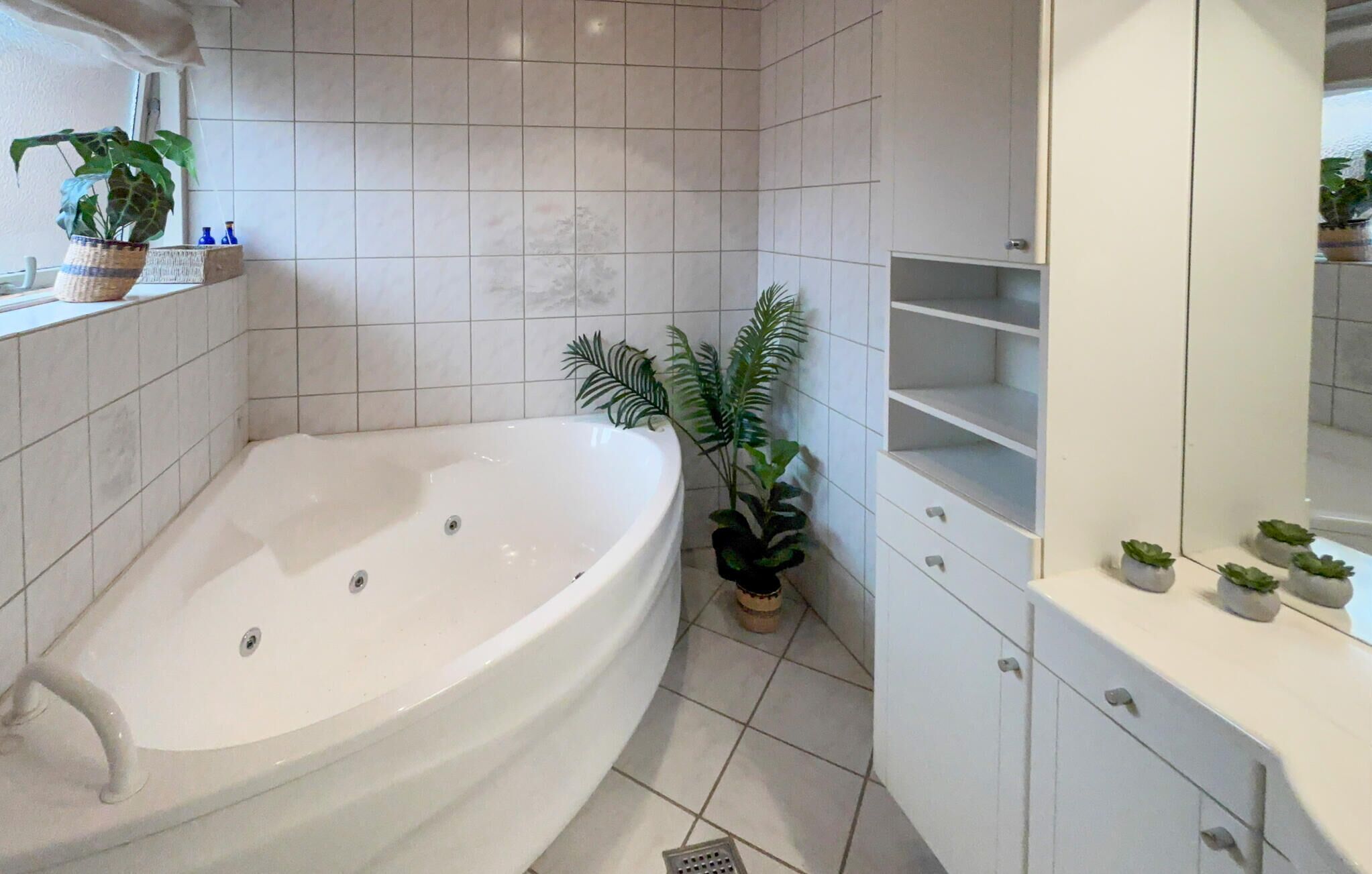 A bathroom with a bathtub, a sink, and a mirror. There are plants and a shelf with toiletries.