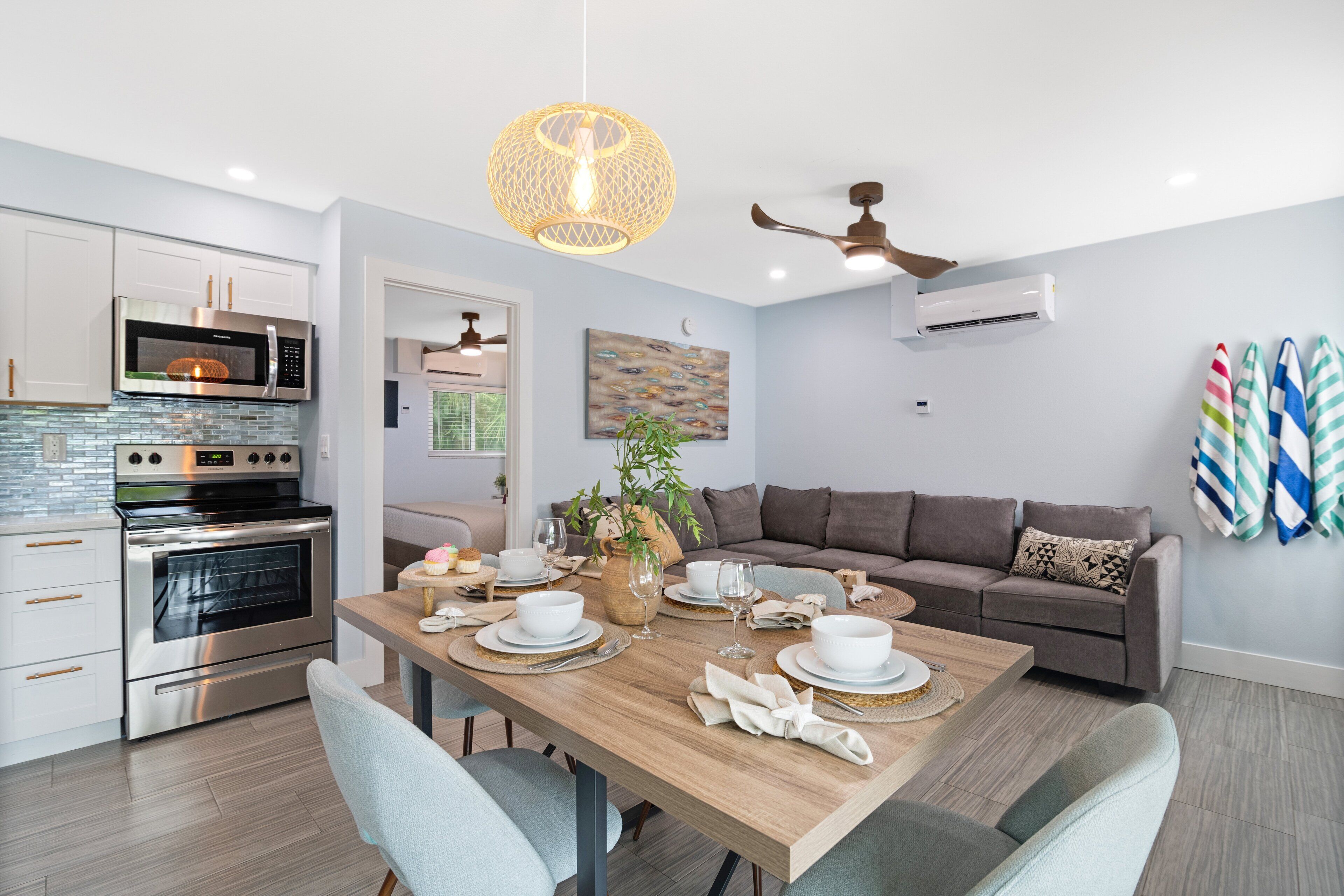 Una cocina moderna con electrodomésticos de acero inoxidable, un comedor con una mesa de madera dispuesta para cuatro personas, y un sofá gris en la sala.