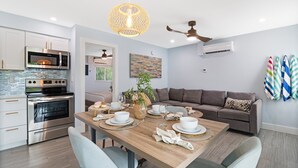 A modern kitchen with stainless steel appliances, a dining area with a wooden table set for four, and a grey sofa in the living area.