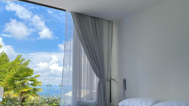 A modern bedroom with a large window, a bed with white and dark blue bedding, and a view of greenery and mountains. - Finca Hotel Los Alpes, 15 minutes from downtown, surrounded by nature. (Manizales)