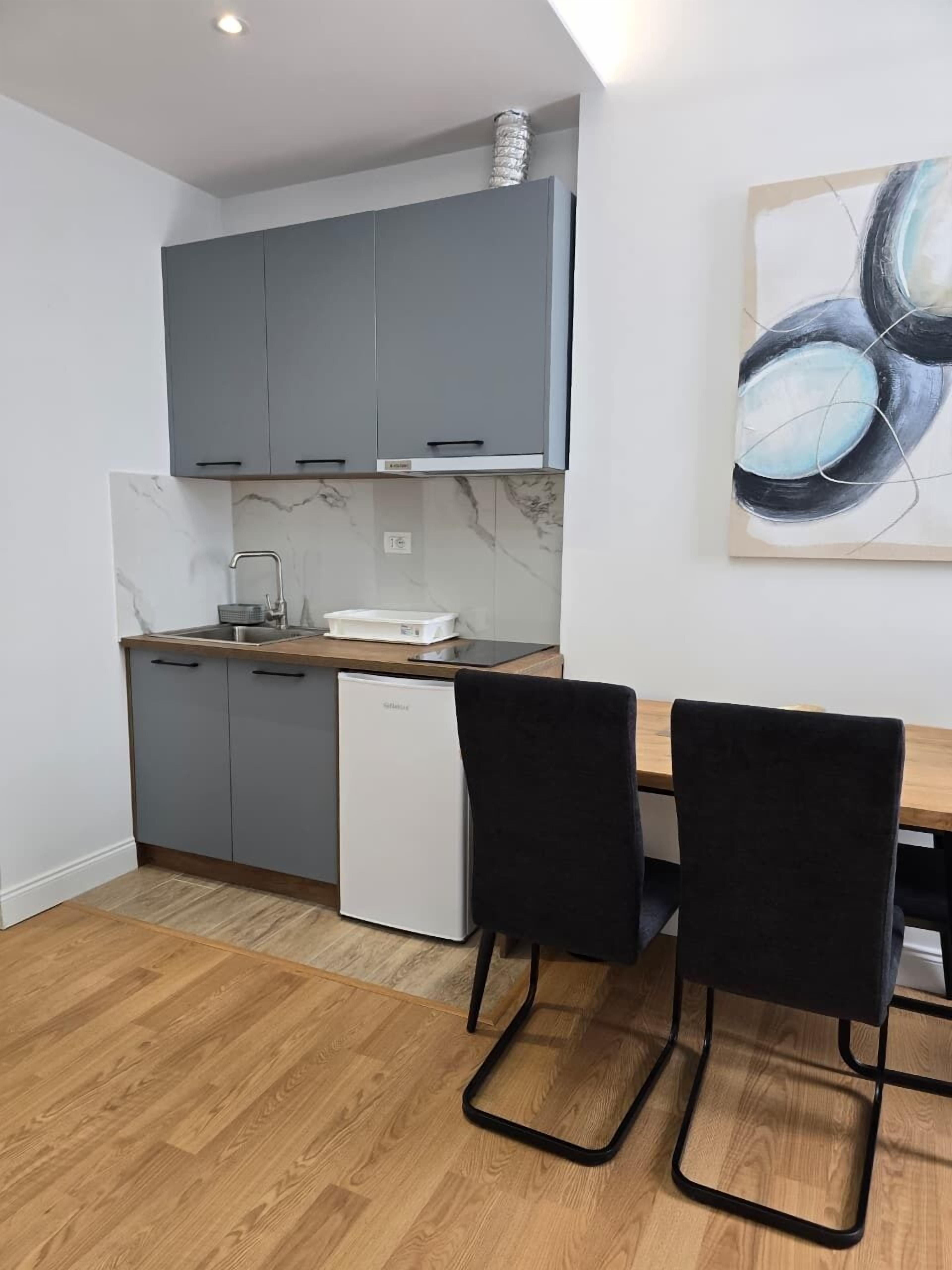 A compact kitchen with grey cabinets, a white dishwasher, and a marble backsplash. There is a dining table with two black chairs and a painting on the wall.
