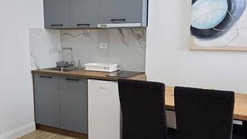 A compact kitchen with grey cabinets, a white dishwasher, and a marble backsplash. There is a dining table with two black chairs and a painting on the wall.