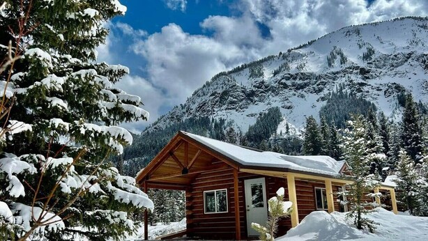 A wooden cabin with a snow-covered roof, surrounded by snow-covered trees and a mountain in the background.