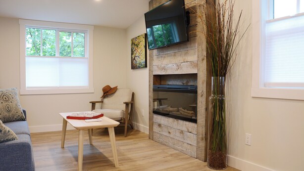Een woonkamer met een bank, een tafel, een aan een houten muur bevestigde televisie en een raam met zonwering.