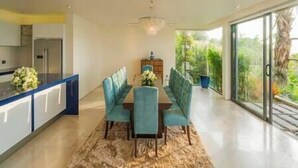 A modern dining area with a blue table and chairs, a kitchen with a refrigerator, and a view of greenery through large windows. - Villa Samira | Kamala Sea View 6BR Villa | Pool, Chef, Spa (Kamala)