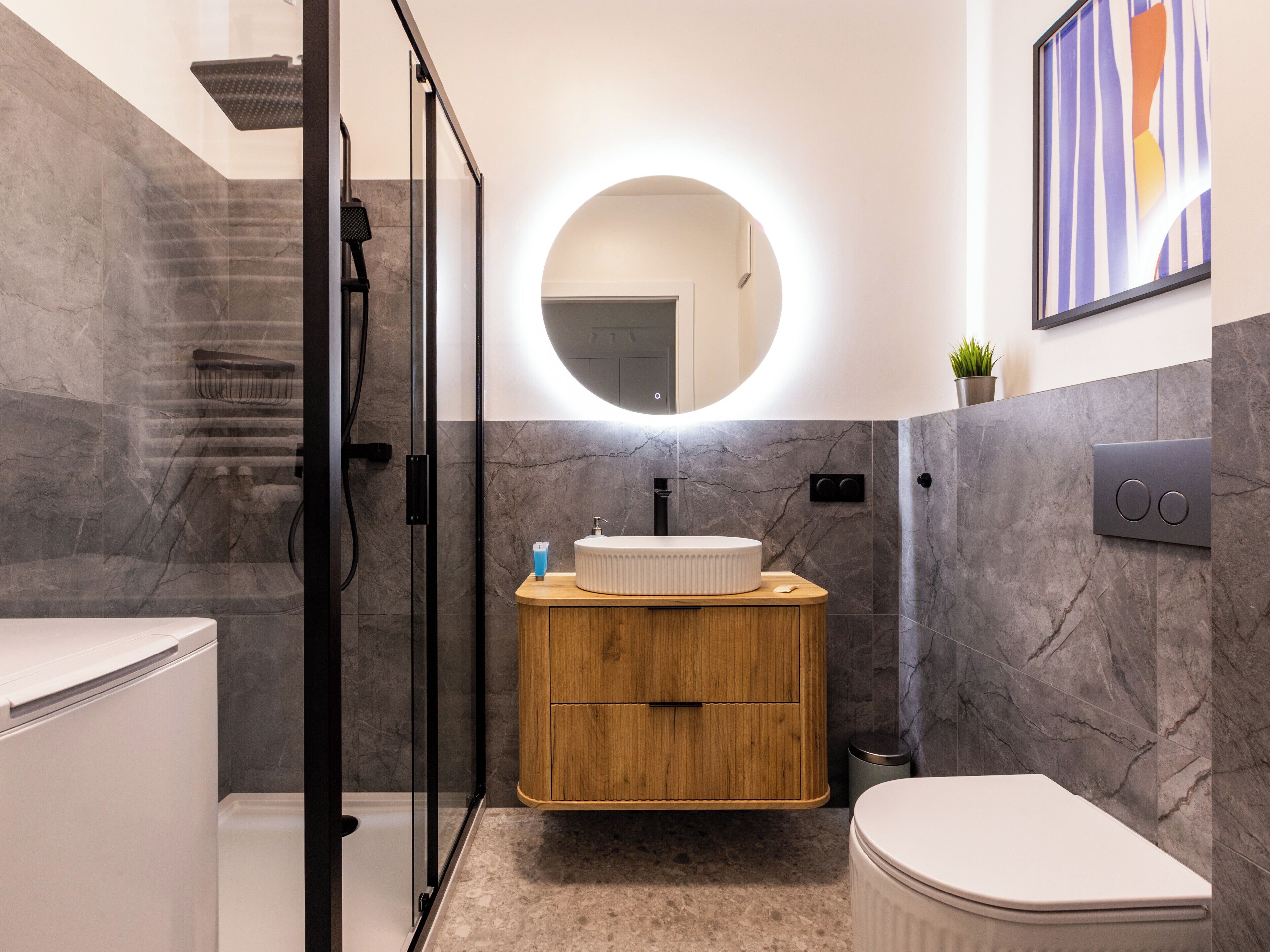Un baño moderno con ducha de vidrio, tocador de madera con un espejo redondo y un inodoro.