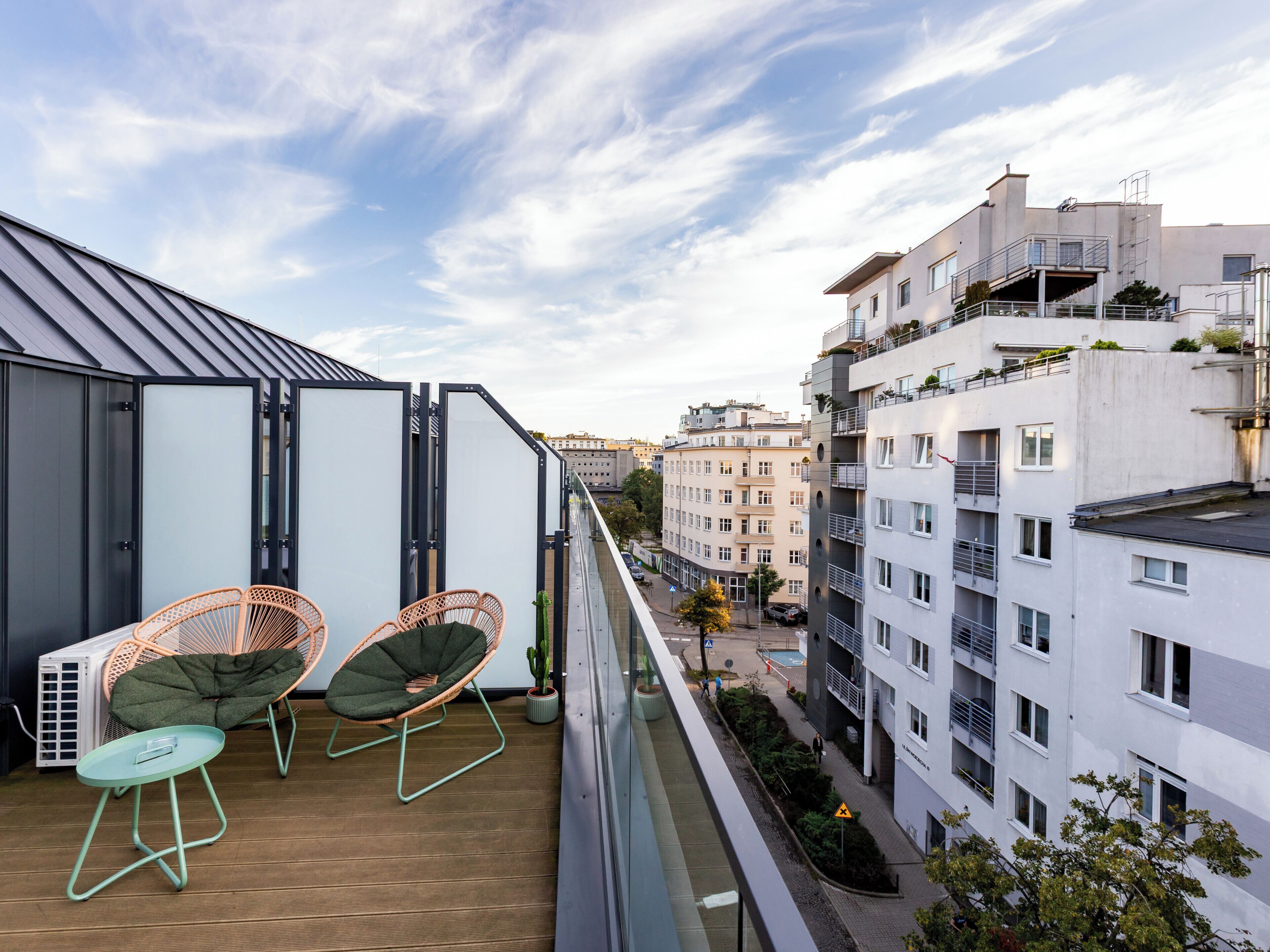 A balcony with two wicker chairs and a small table, overlooking a city street with buildings and a clear sky.