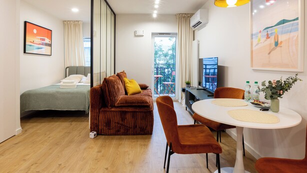 Habitación de hotel moderna con cama, sofá, mesa de comedor y sillas. Cuenta con un ventanal con cortinas, un equipo de aire acondicionado y un cuadro en la pared.