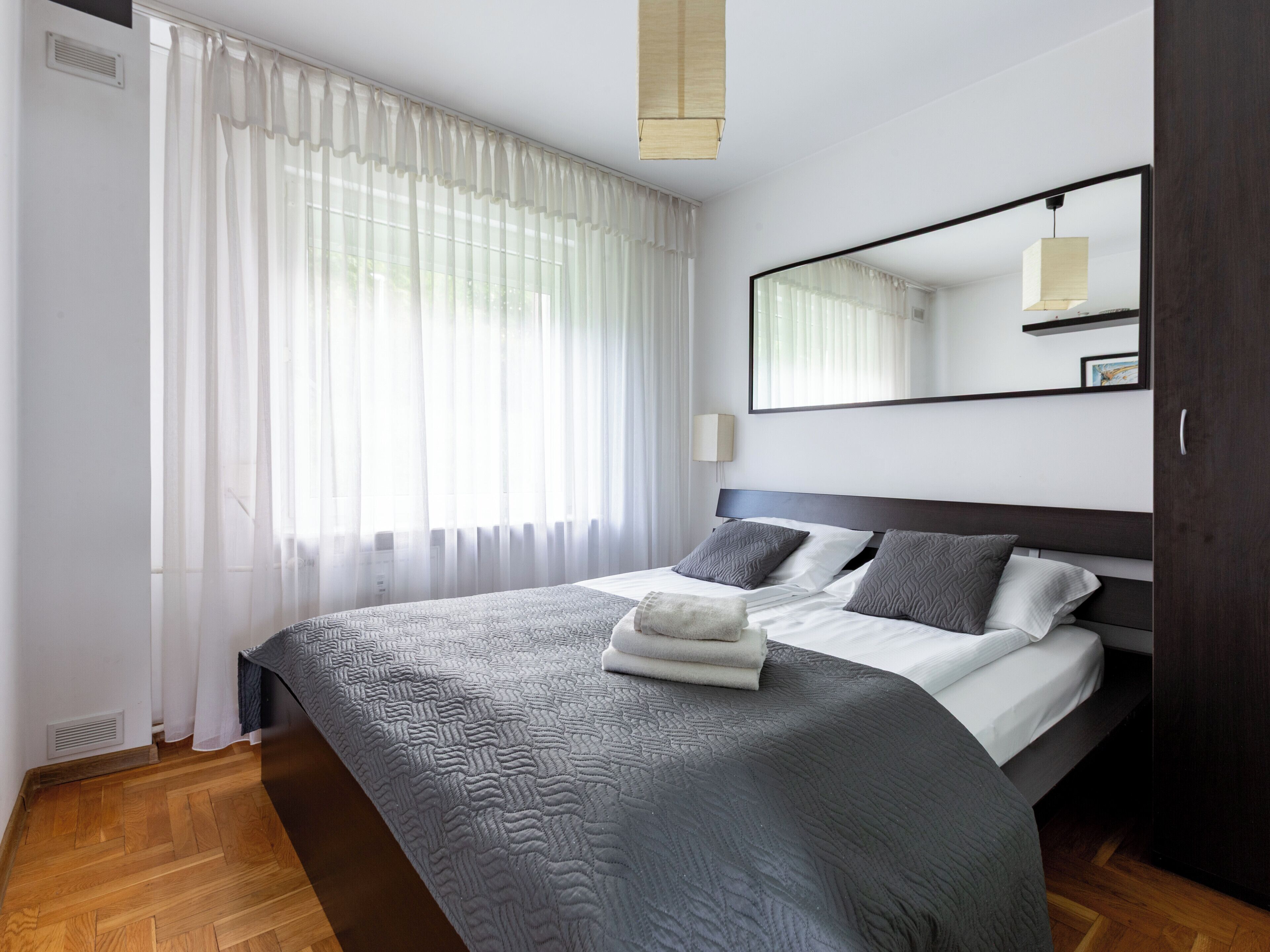 Quarto de hotel moderno com uma cama grande, piso de madeira e uma janela ampla com cortinas leves.