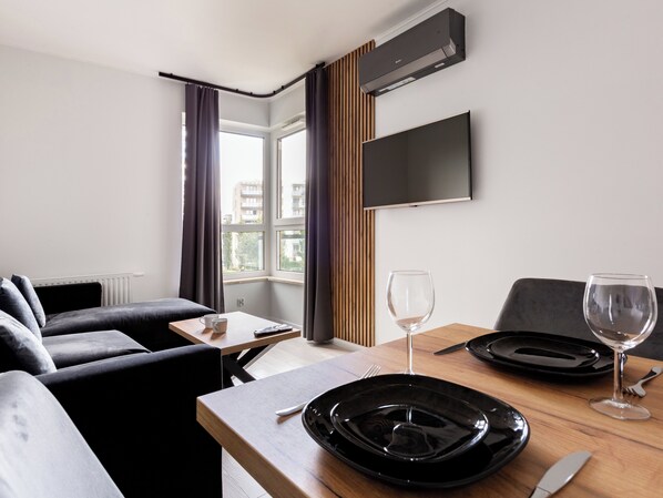A modern hotel room with a sofa, a dining table set with plates, glasses, and cutlery, a flat-screen TV mounted on the wall, and a window with curtains.
