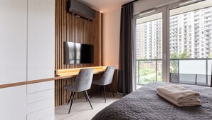 Een moderne hotelkamer met een groot bed, een bureau met twee stoelen, een flatscreen televisie bevestigd aan een houten paneel, en een balkon met uitzicht over de stad.