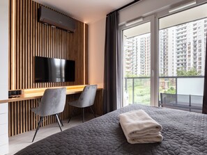 A modern bedroom with a large bed, a desk with two chairs, a flat-screen TV, and a balcony with a city view.