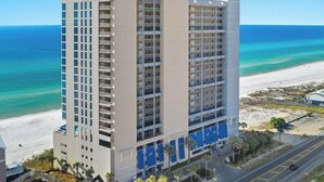 A high-rise hotel with multiple balconies, a beach view, and a parking area with palm trees.