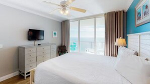 A hotel room with a large bed, a television on a dresser, a balcony with curtains, and a ceiling fan.