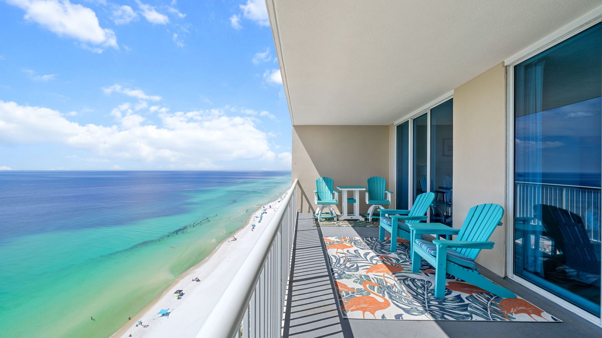 Un balcone con vista sull'oceano, acqua turchese e una spiaggia con persone. Si vedono sedie e un tavolo turchesi.