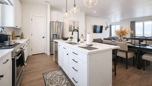 Une cuisine moderne avec des meubles blancs, un îlot central, des appareils en acier inoxydable, un éclairage suspendu et un coin repas offrant une vue agréable.