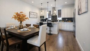 Une cuisine moderne avec des meubles blancs, un îlot central, des luminaires suspendus et un coin repas avec une table dressée pour quatre personnes.