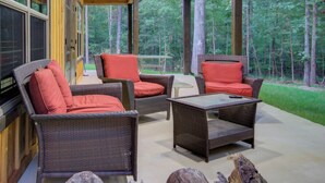 A covered outdoor seating area with wicker furniture and red cushions, overlooking a forested area.