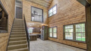 A spacious log cabin interior with a central kitchen, a staircase leading to a loft, and large windows offering views of the outdoors.