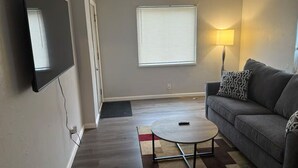 Un salón con un televisor de pantalla plana montado en la pared, un sofá gris con cojines estampados, una mesa de centro redonda y una lámpara de pie.