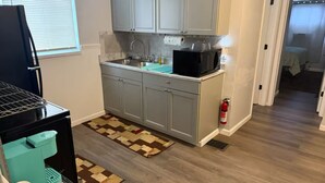 Cocina compacta con gabinetes grises, un microondas negro y un lavamanos. Hay un extintor cerca de la puerta y una alfombra pequeña en el suelo.