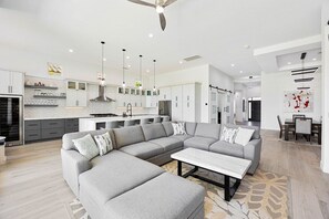 A modern living space with a grey sectional sofa, a white coffee table, and a kitchen area with white cabinets and pendant lighting. - Mountain View Oasis | Pool, Spa, Golf, EV Charger! (La Quinta)