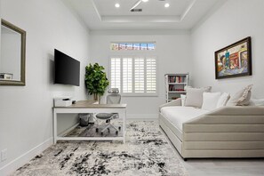 A modern bedroom with a bed, a desk, a chair, a TV, a bookshelf, and a window. - Mountain View Oasis | Pool, Spa, Golf, EV Charger! (La Quinta)