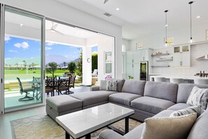 A modern living room with a sectional sofa, a glass door leading to a patio, and a kitchen with white cabinets and pendant lighting. - Mountain View Oasis | Pool, Spa, Golf, EV Charger! (La Quinta)