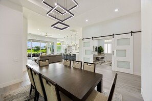 A modern dining area with a large table and chairs, a kitchen in the background, and a sliding glass door leading to a patio.