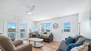 A spacious living room with a gray sofa, a beige recliner, and a coffee table. There are two windows with views of a house and palm trees. - 200 Steps To Beach, Balcony Gulf Views, Firepit, Dog Friendly, 3BR Beach Home (Gulf Shores)