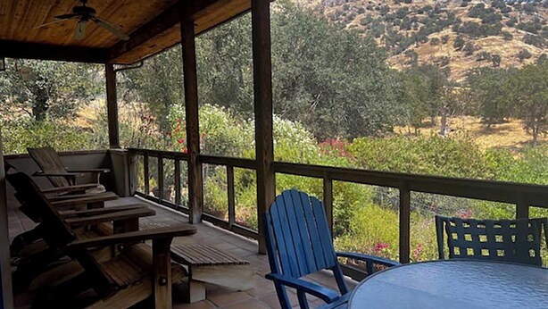A covered patio with wooden furniture, including a table and chairs, overlooking a hilly landscape with trees and shrubs. - Heidi's Artistic Abode, peaceful relaxation! (Three Rivers)