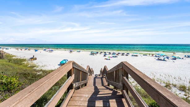 Eine Holztreppe führt zu einem Strand mit Sonnenschirmen und Menschen.