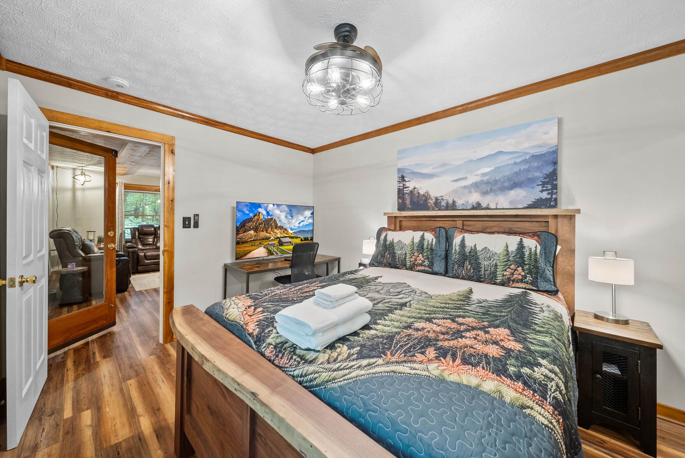 A bedroom with a bed, a desk, a chair, a painting, and a ceiling fan.