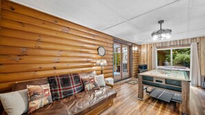 Une pièce chaleureuse avec un mur en bois brut, un canapé en cuir marron agrémenté de coussins décoratifs, une table recouverte d’une nappe verte et un ventilateur de plafond.